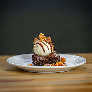 A plate with a slice of brownie and a scoop of ice cream on top with sauce and biscuit crumbled over it.